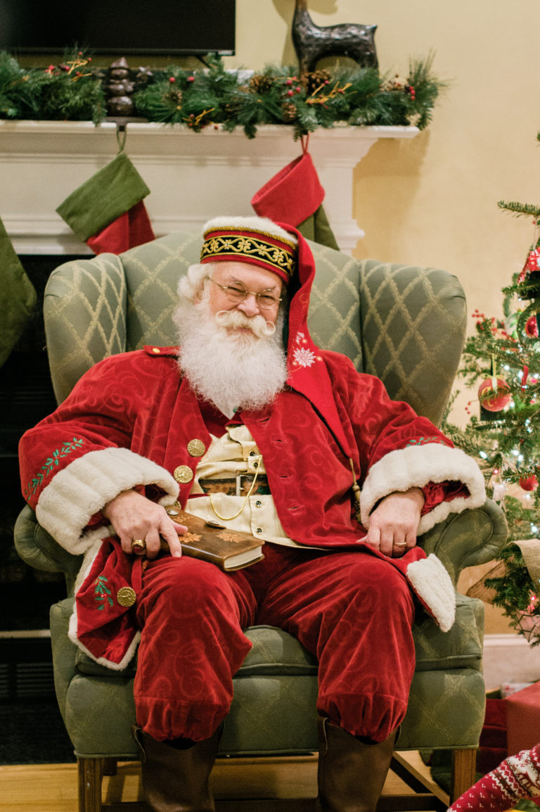 A man dressed as Santa Claus sits in an easy chair in front of a fireplace of stockings.