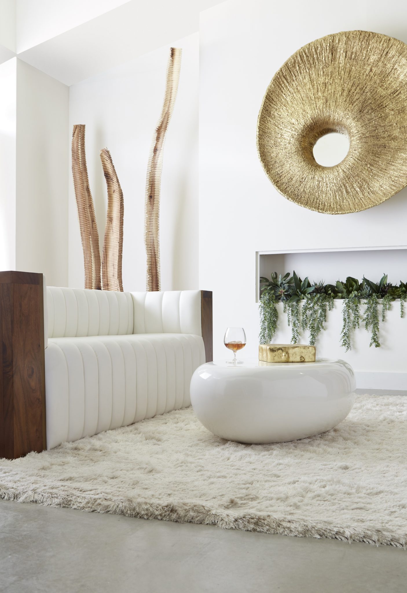 A room with a wood couch sitting on a fuzzy carpet. A coffee table with a wine glass sits in front of some plants and under a modern mirror.