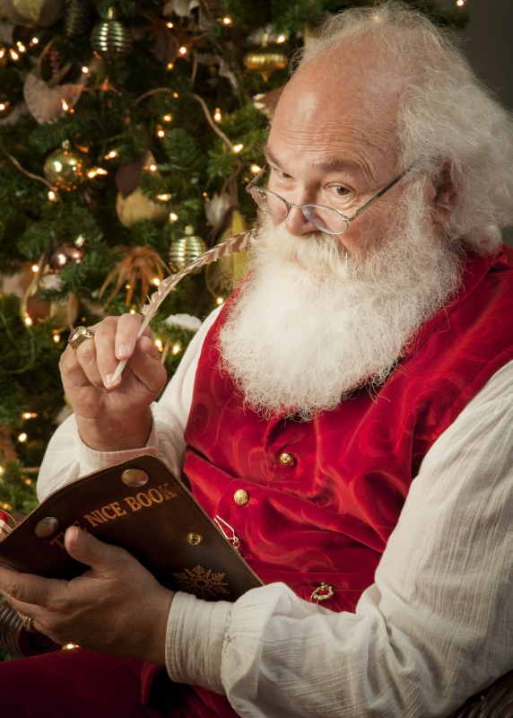 Santa Cliff, a man dressed as Santa Claus, sits in front of a tree looking in a book.