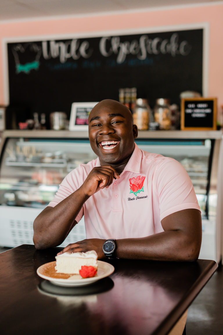 Daniel Gray sits at a table with cheesecake on it, wearing a shirt that says "Uncle Cheesecake" in front of a blackboard with a menu that says "Uncle Cheesecake."