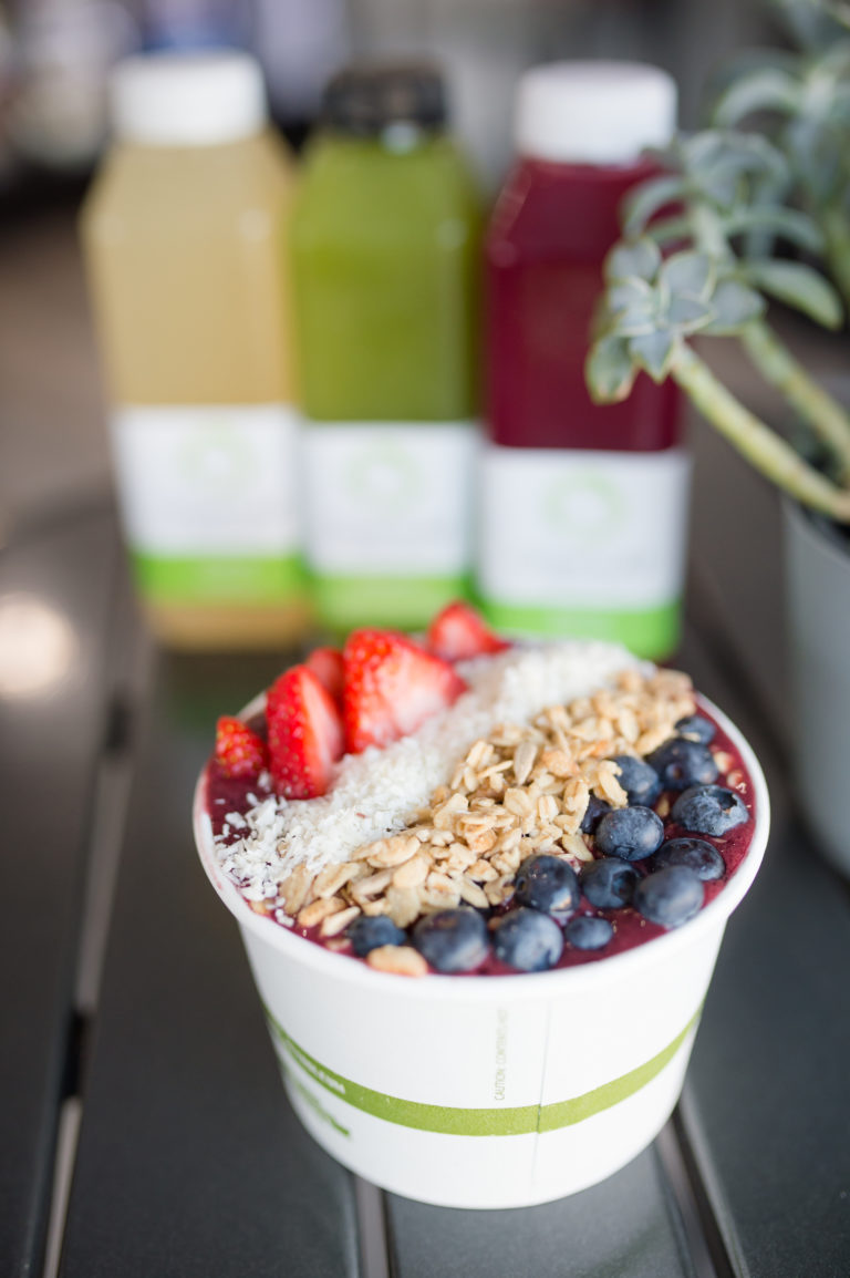 A smoothie bowl of blueberries, oatmeal, coconut, and strawberries sits in front of three bottles of pressed juice at Organic AF.