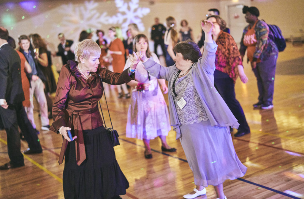 Two women dance together in the gym, while other prom guests watch.
