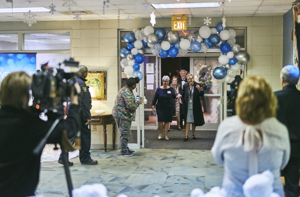 Two women walk under a blue and silver balloon arch into a church as people on the sidelines clap and smile.
