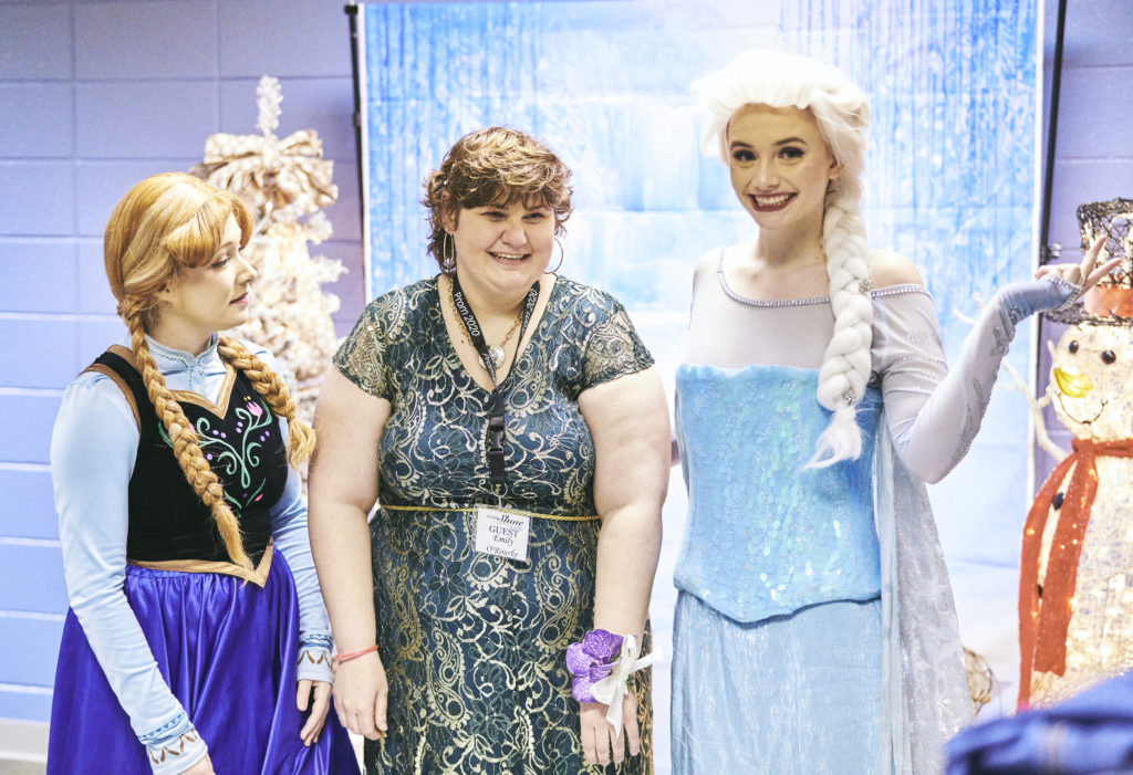 A girl in a dress stands between two actresses dressed as Princess Elsa and Princess Ana from Disney's Frozen.