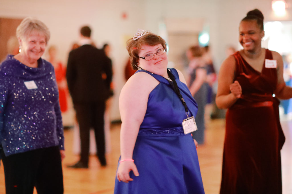 A girl dances in a blue prom dress while another girl and an older woman watch and smile.