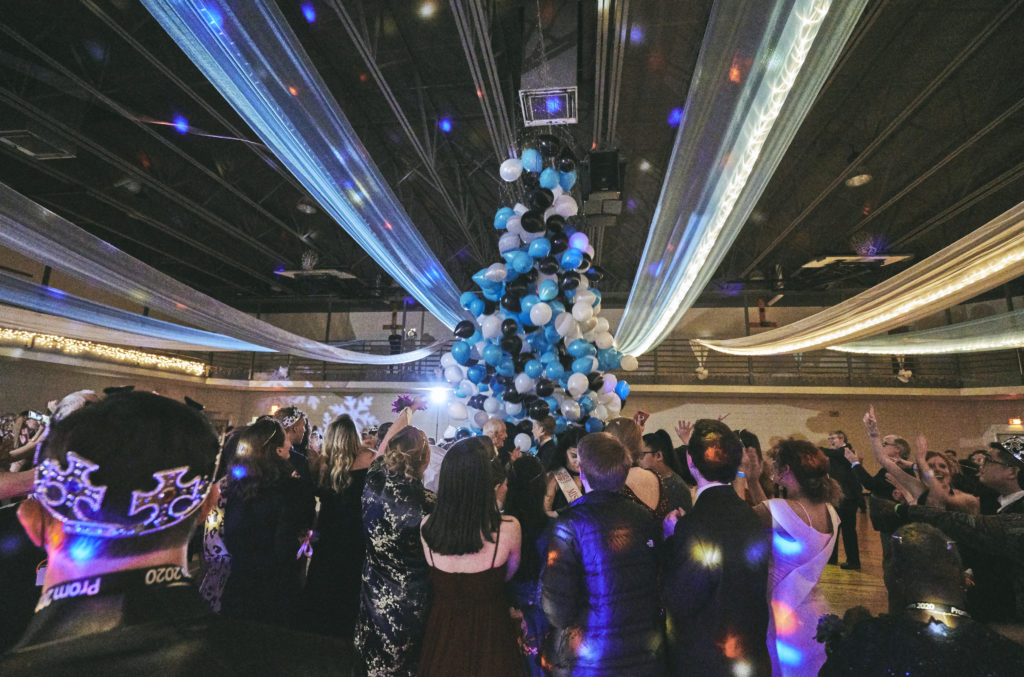 A dance floor with bright lights and a balloon sculpture of black and blue and white balloons. Groups of party goers dance under the balloons.