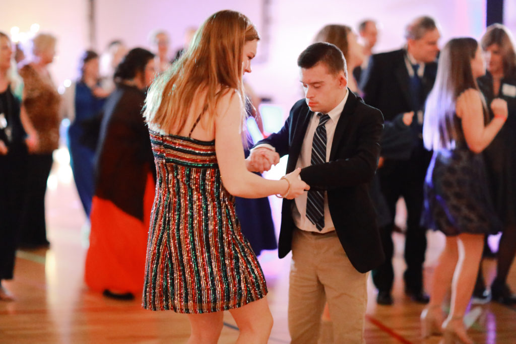 A girl and a boy in dressy clothes dance together on a gym floor, as others dance in the background.