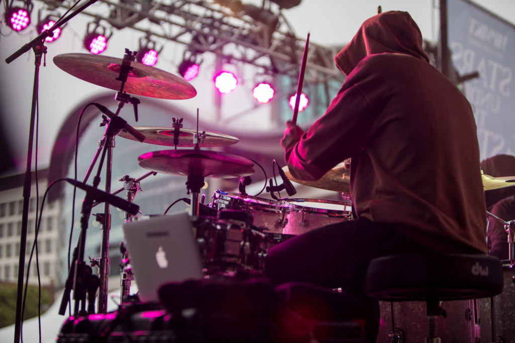 A drummer wearing a hoodie plays drums under pink concert lights.