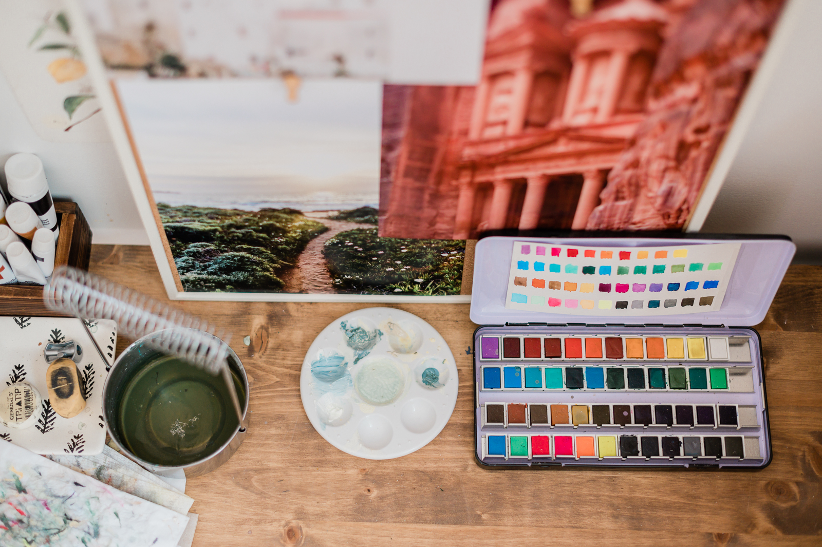 A desk has a palette of paints on it, with artwork in the background.