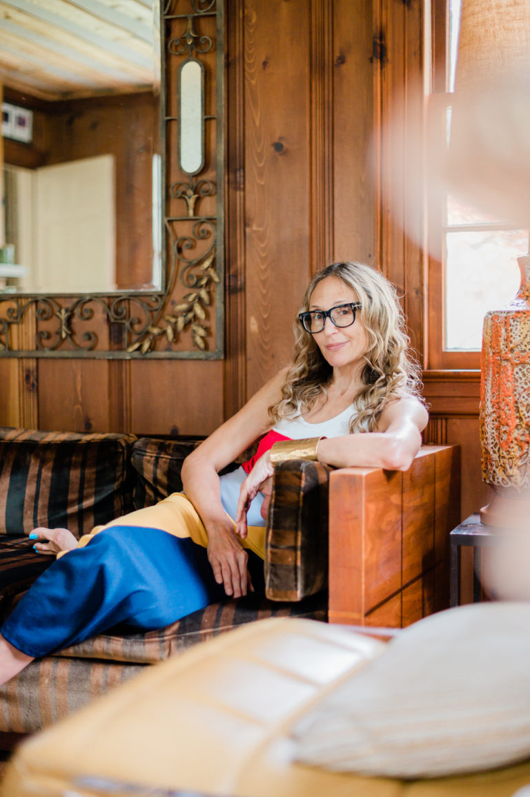 A woman in big glasses and a bright dress lounges on a wooden, cushioned sofa..