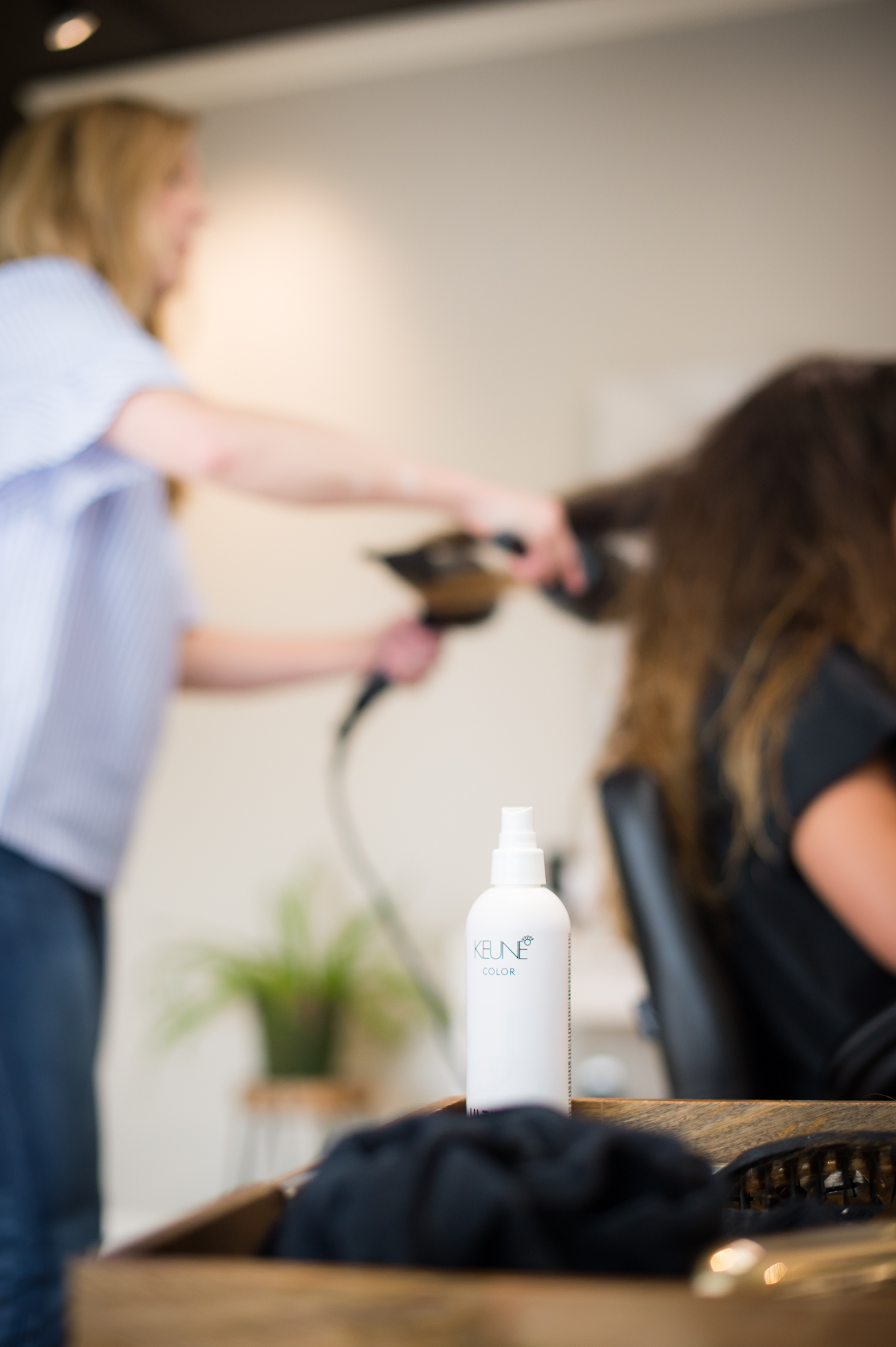 Brittany Hall blow dries a woman's hair out of focus with a bottle of hair styling product in the foreground.