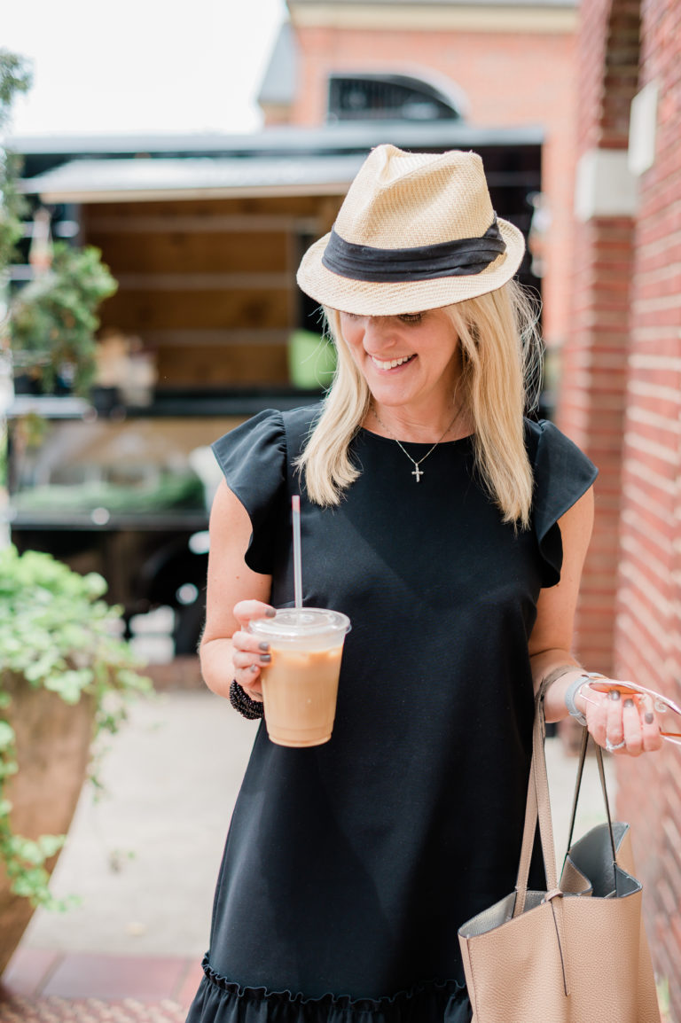 A woman in a black dress wearing a straw and black hat smiling and holding an iced coffee.