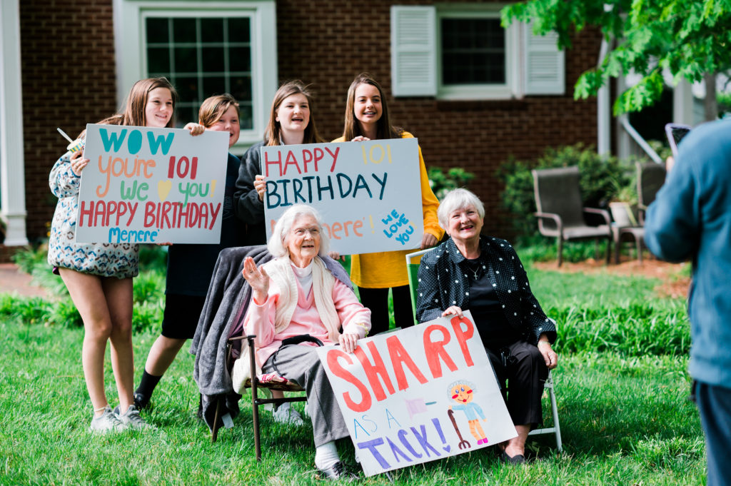A group of young girls holding birthday signs stand behind Jean and her daughter seated on the front lawn.