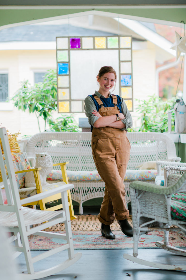 Annabella Boatwright, owner of Neon Tumbleweed Studio, stands in overalls on a porch.