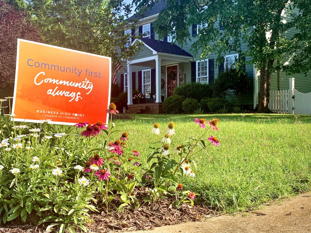 A yard sign that says "Community First. Community Always." sits in a front yard among flowers.