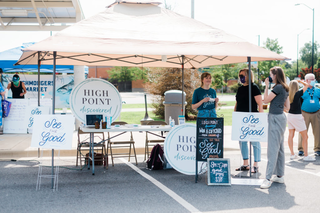 People stand at a table at the High Point Farmer's Market that has signs saying "High Point Discovered" and "See the Good."
