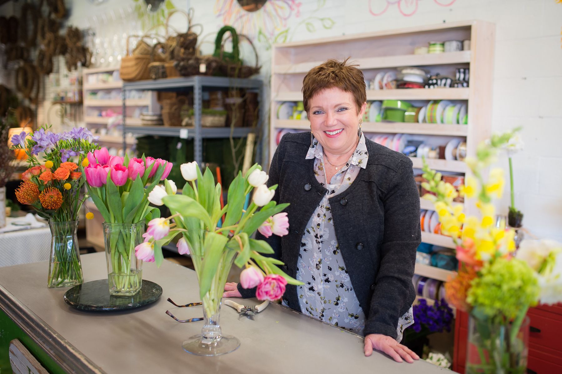 Sami Price, owner of Just Priceless, stands smiling at a counter with several vases of cut flowers.