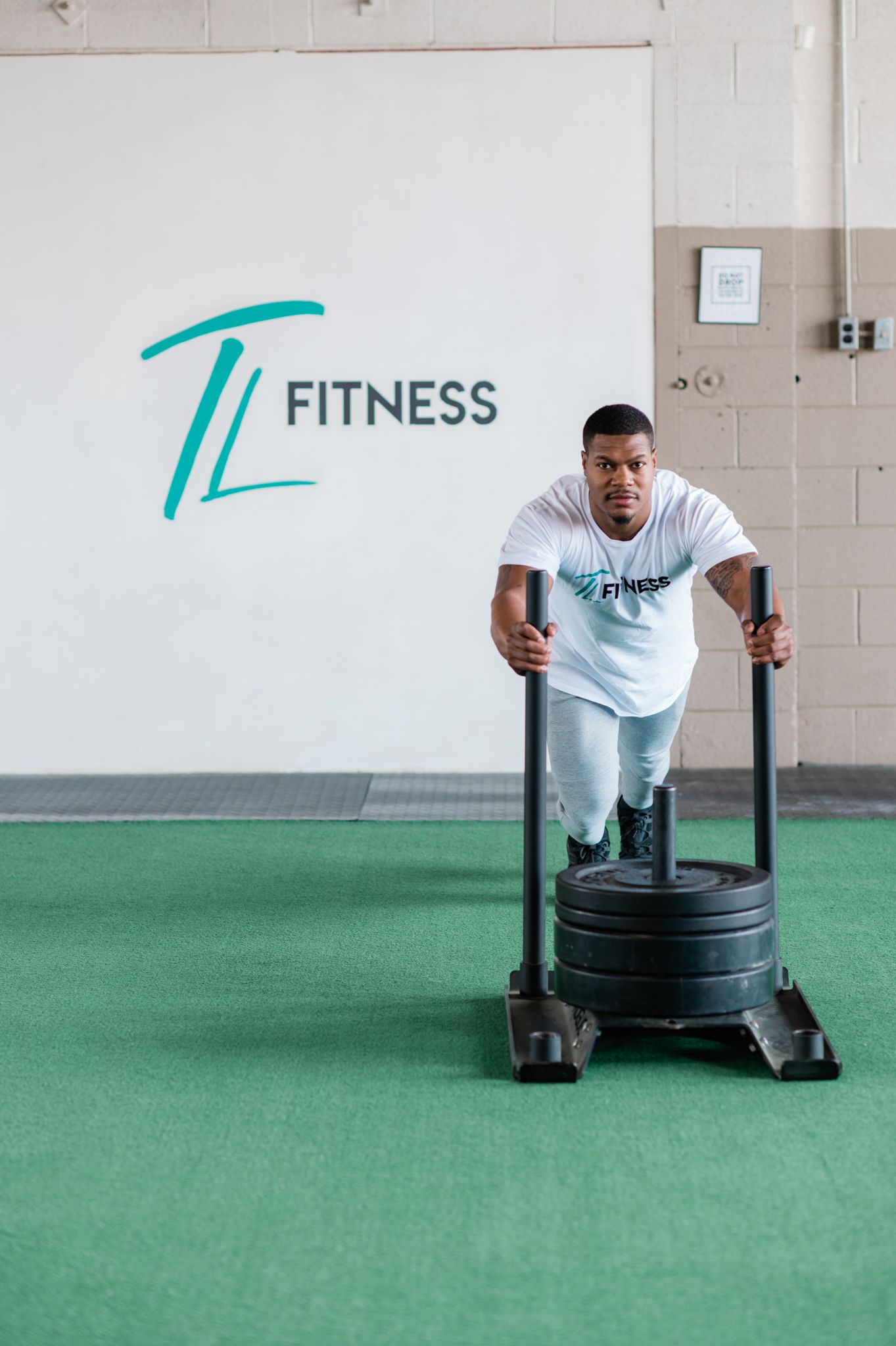 Man looks at camera while pushing weights on a lift forward. In the background there is a wall that reads "TL Fitness."