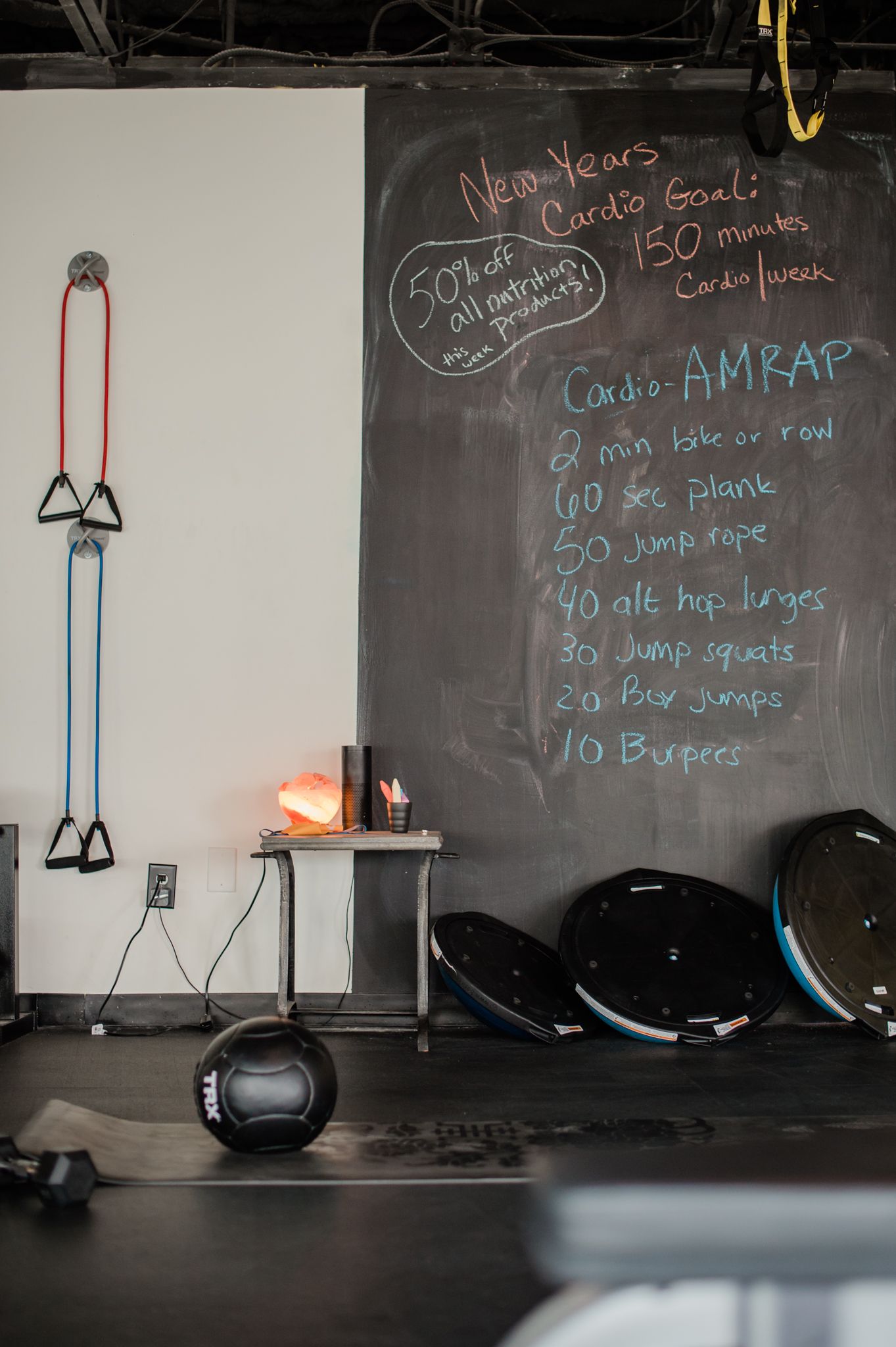 A gym with a chalkboard with exercise rep instructions written on it. Around the chalkboard are several barbell weights, a medicine ball, and other exercise equipment.