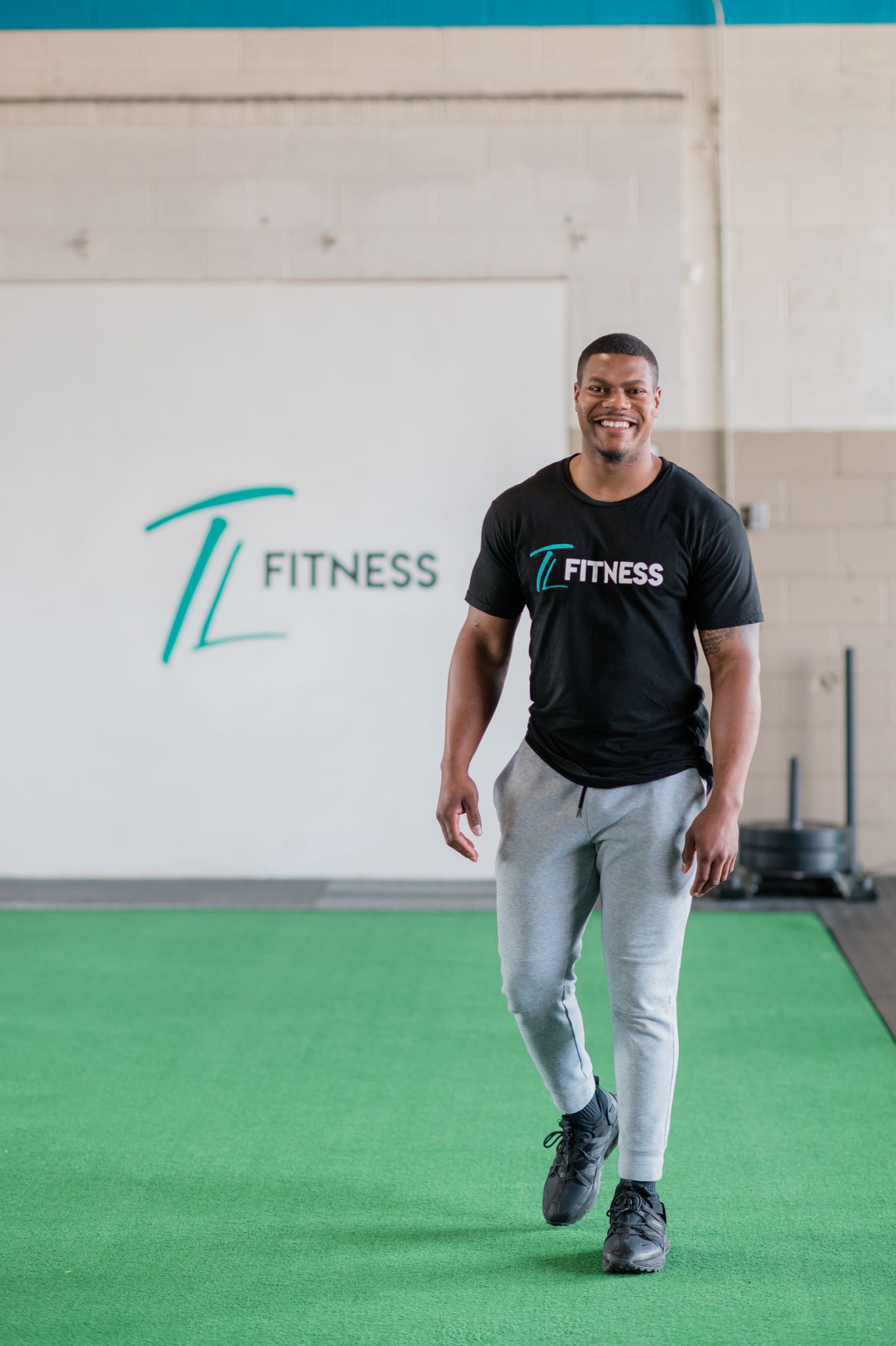 A man, Torrey Lowe, smiles and walks towards the camera. He is in a gym with "TL Fitness" written on the wall.