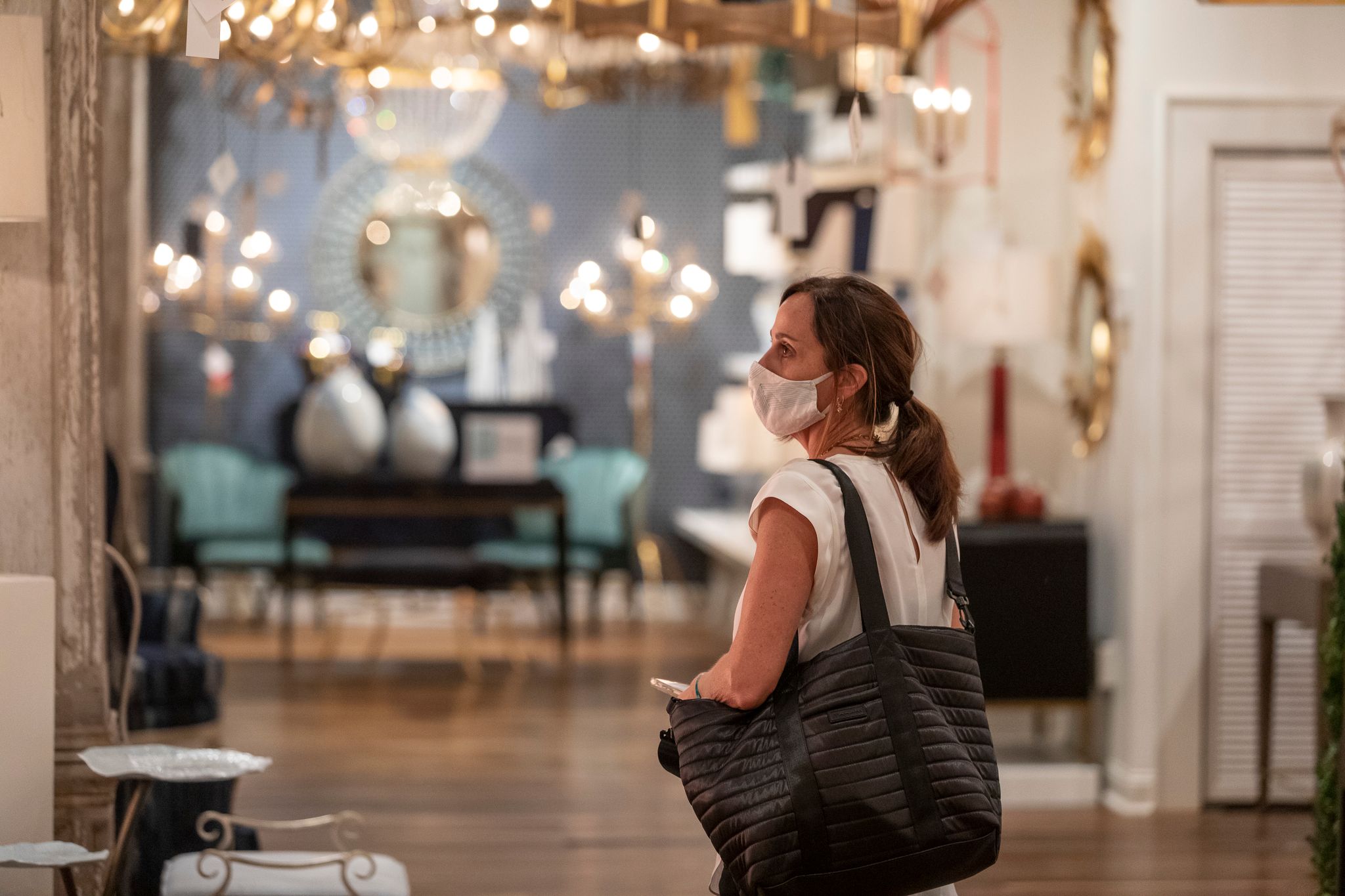A woman stands in a furniture showroom looking at various objects. She is carrying a large bag and wearing a cloth mask.