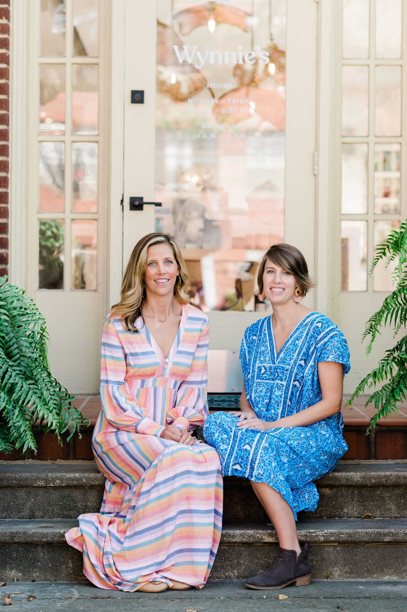 Two women sit side by side on the front steps of a boutique storefront, smiling at the camera.