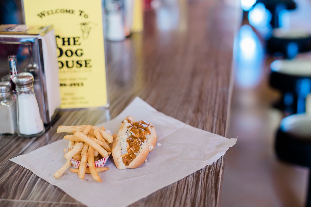 A counter at a restaurant with a hotdog with chili and french fries lies on a piece of butcher paper in front of a yellow menu that says "The Dog House."