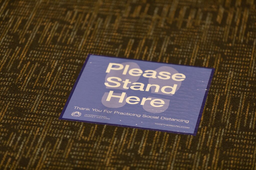 On gray carpet lays a sign with two footprint outlines that says, "Please Stand Here."