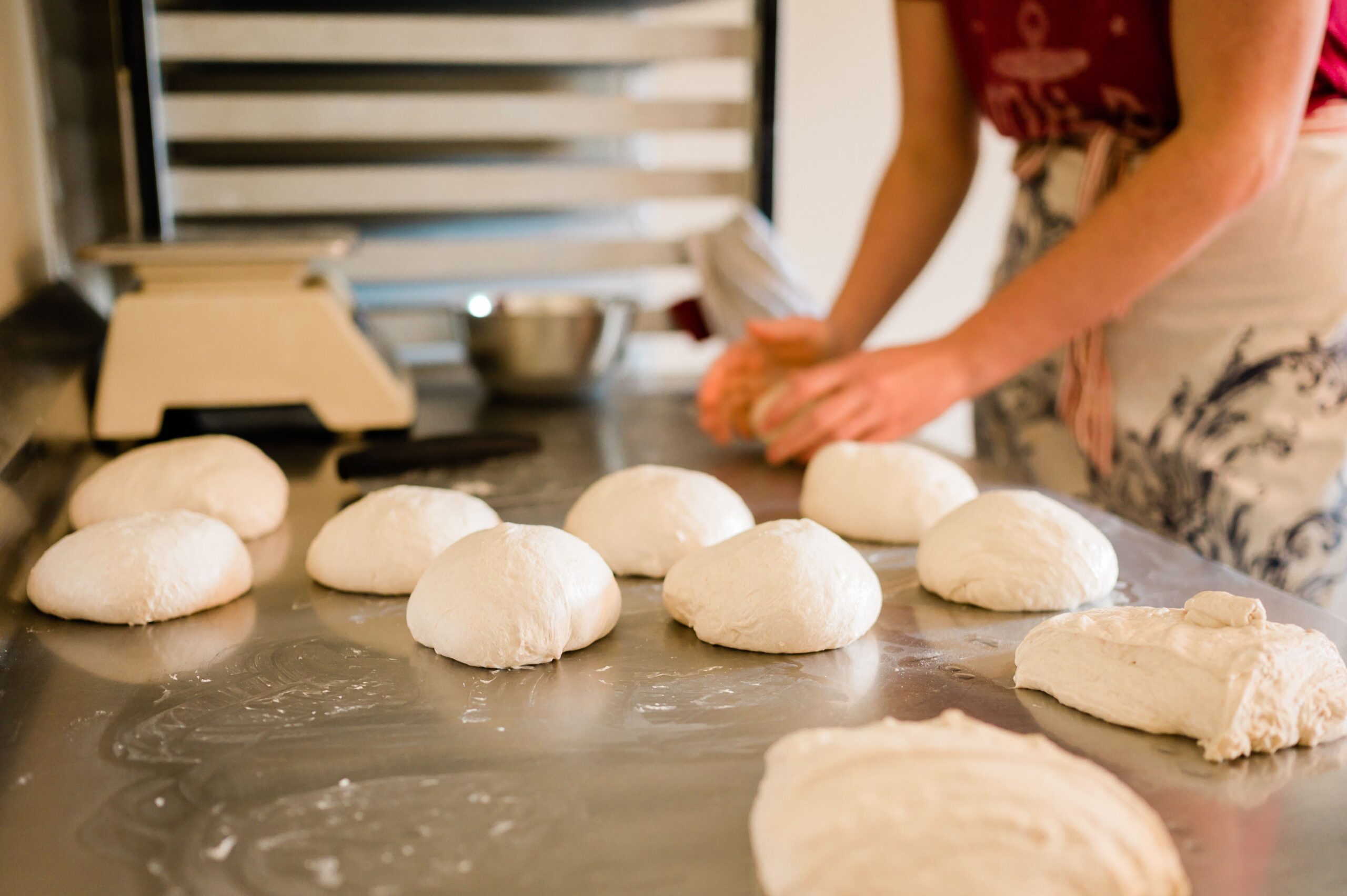 A steel counter with rolls of uncooked bread dough stand while a woman's hands are shown kneading the dough.