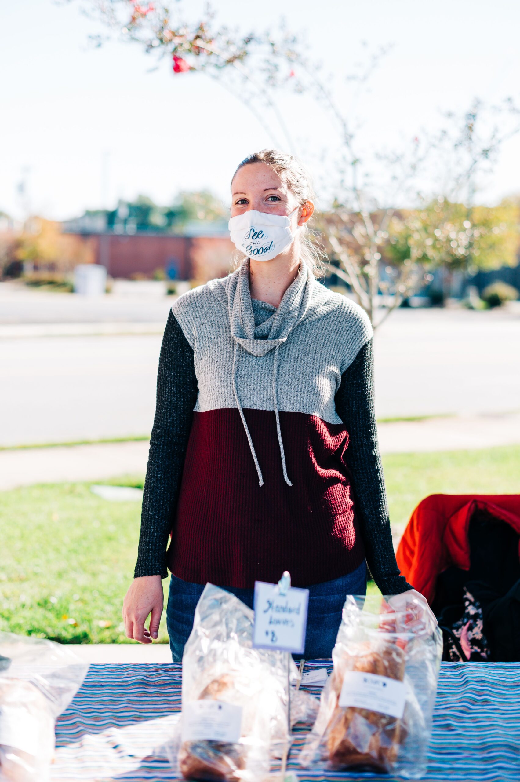 Woman stands outside by a table with wrapped loaves of bread on it. She is wearing a sweatshirt and a fabric face mask that says, "See the Good."