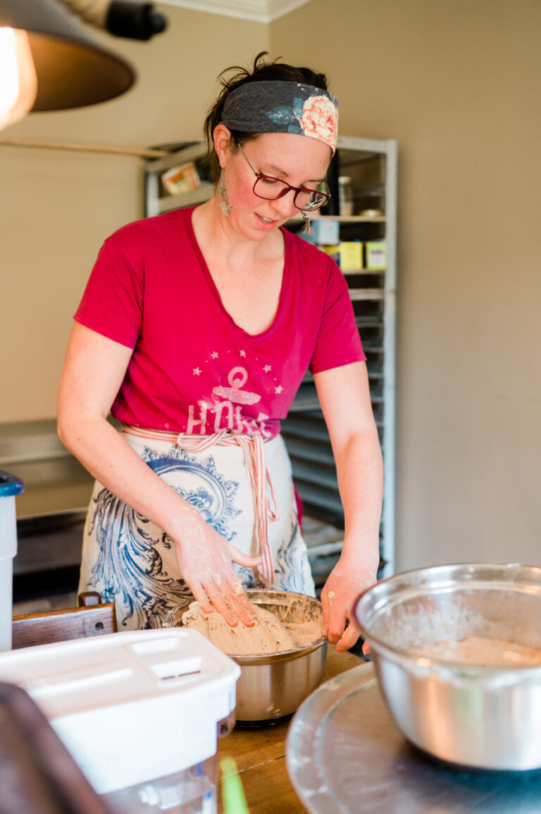 A woman in a red t-shirt and apron stands in a kitchen kneading a large lump of bread dough.