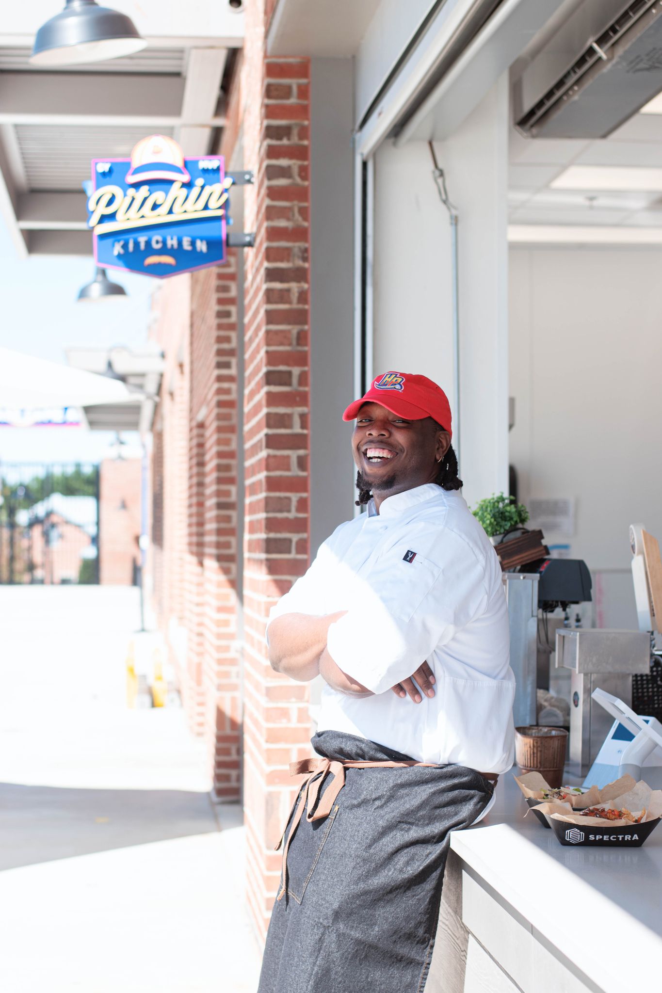 Man in white chef's jacket and red baseball hat leans against outdoor kitchen counter with a "Pitchin' Kitchen" sign hanging above him.