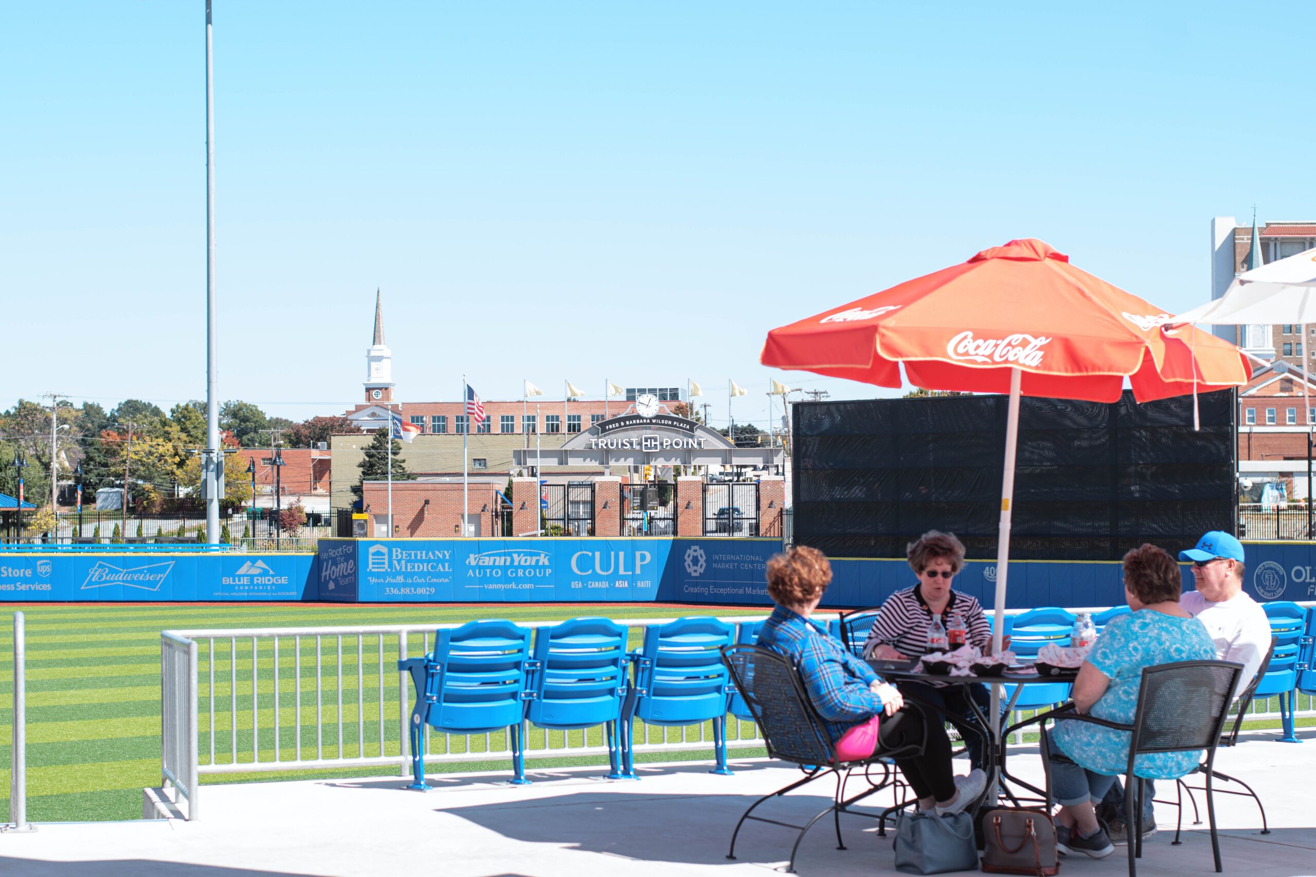A group of four people sit around an outdoor table above the High Point Rockers' baseball field under a red umbrella.