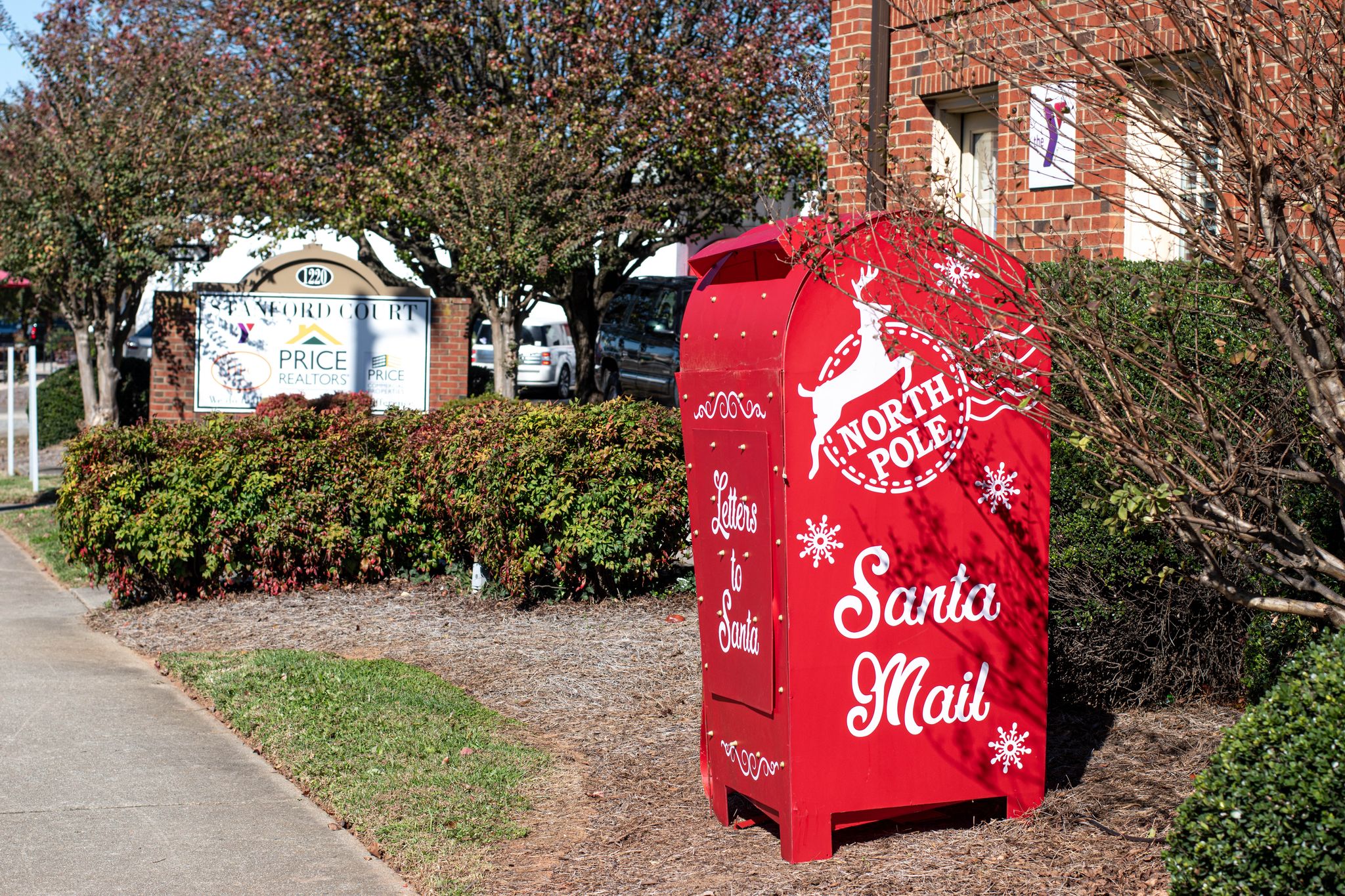 A red metal mailbox that says "North Pole: Santa Mail" on it, sitting in a patch of grass in front of Price Realtors.