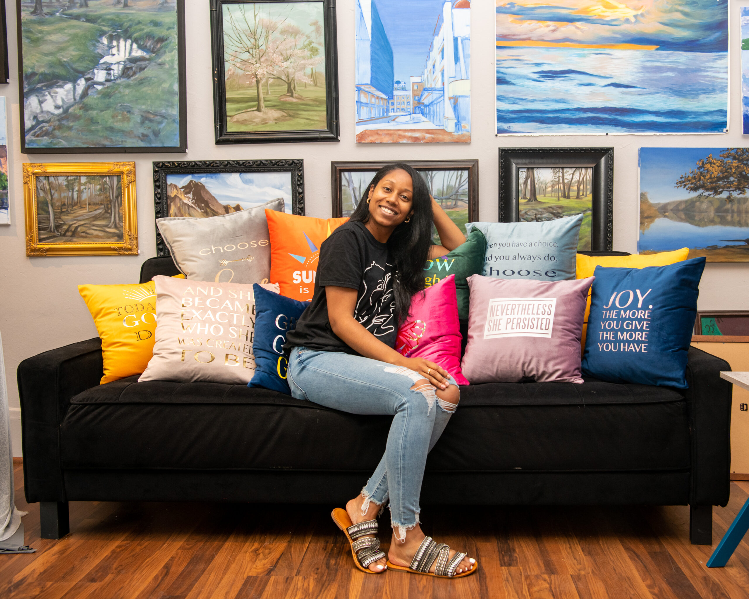 Maya Graham, curator for the Art Market exhibit at the Gallery on Main sits on a sofa with pillows in front of a wall of paintings in High Point, NC.