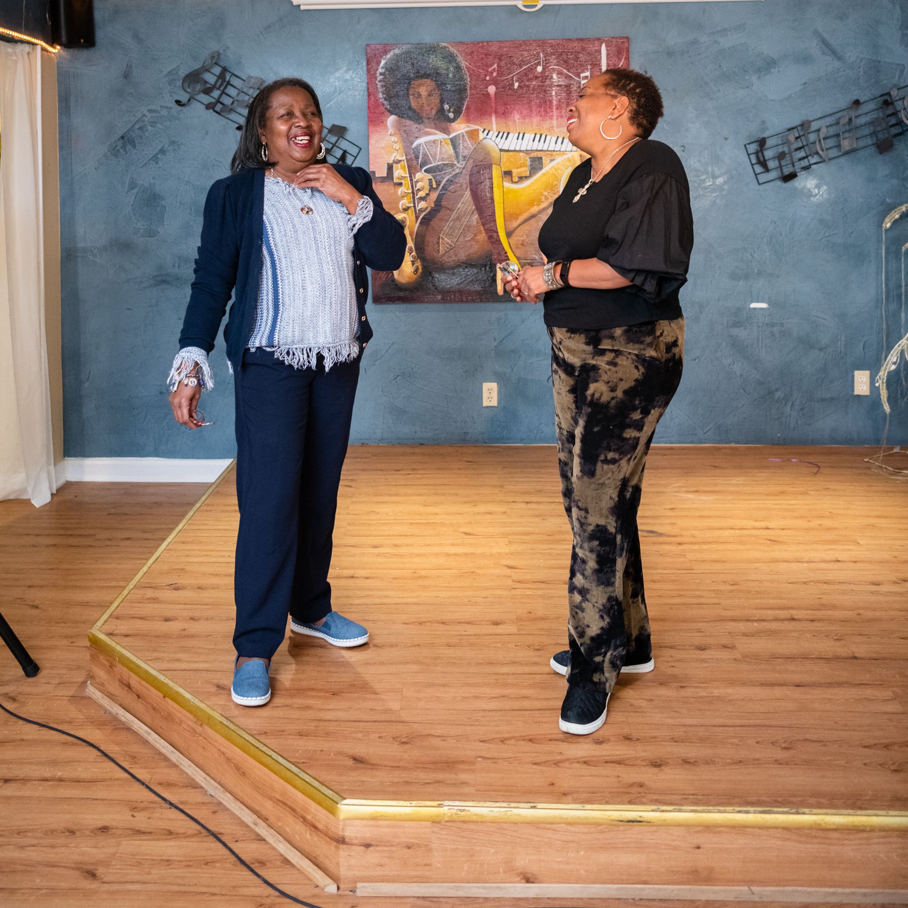 Raynetta Jackson and Rishaunda Moses stand in front of artwork by Anthony Belton on the stage at Jackie's Place in High Point, NC.