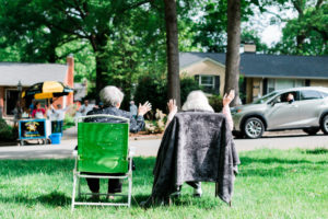 A shot from behind of Jean and her daughter sitting in chairs in their yard, waving to passerbys in cars.