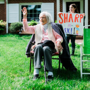 A young girl holds a hand-drawn sign that says, "Sharp as a Tack" in the foreground, with Jean and her daughter seated in the background in High Point, NC