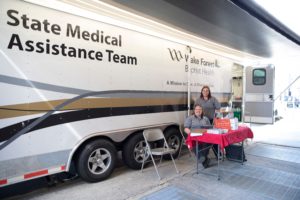 A trailer reading "State Medical Assistance Team" is parked by a curb. A woman sits at a fold out table in front of the trailer and another woman stands beside it.